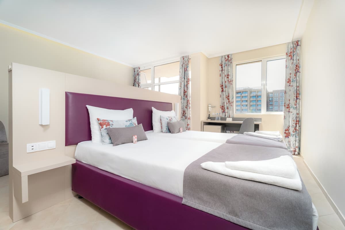 A modern hotel room with a large bed, a desk, and a view of buildings through the window.