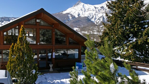 A wooden cabin with large windows, a bench outside, and a sign pointing to the right. - VVF Les Ecrins Champsaur, Saint Bonnet en Champsaur (Saint-Bonnet-en-Champsaur)