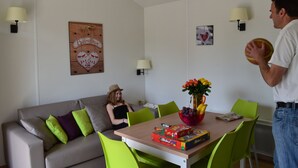 A living room with a sofa, a dining table with chairs, a lamp, and a wall with a welcome sign. - VVF Les Ecrins Champsaur, Saint Bonnet en Champsaur (Saint-Bonnet-en-Champsaur)