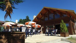 An outdoor seating area with tables and chairs, umbrellas, and a wooden building with large windows. - VVF Les Ecrins Champsaur, Saint Bonnet en Champsaur (Saint-Bonnet-en-Champsaur)