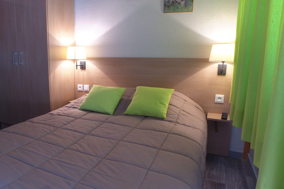 Ein ordentlich bezogenes Bett mit Kopfteil, zwei Nachttischlampen und einem grünen Vorhang.