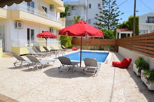 Área da piscina com espreguiçadeiras e guarda-sóis vermelhos, cercada por edifícios e uma cerca de madeira.
