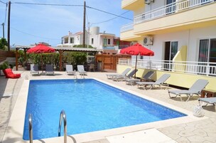 Piscina retangular com escada de metal, rodeada por espreguiçadeiras e guarda-sóis vermelhos.