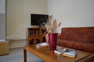 Una sala de estar con una mesa de centro de madera, un jarrón rojo con plantas secas, una estantería y un televisor de pantalla plana montado en la pared.
