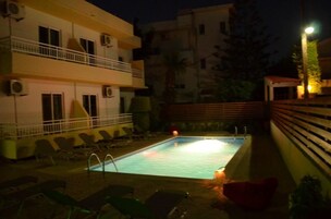 Una piscina con sillones de descanso y una cerca, rodeada de edificios durante la noche.