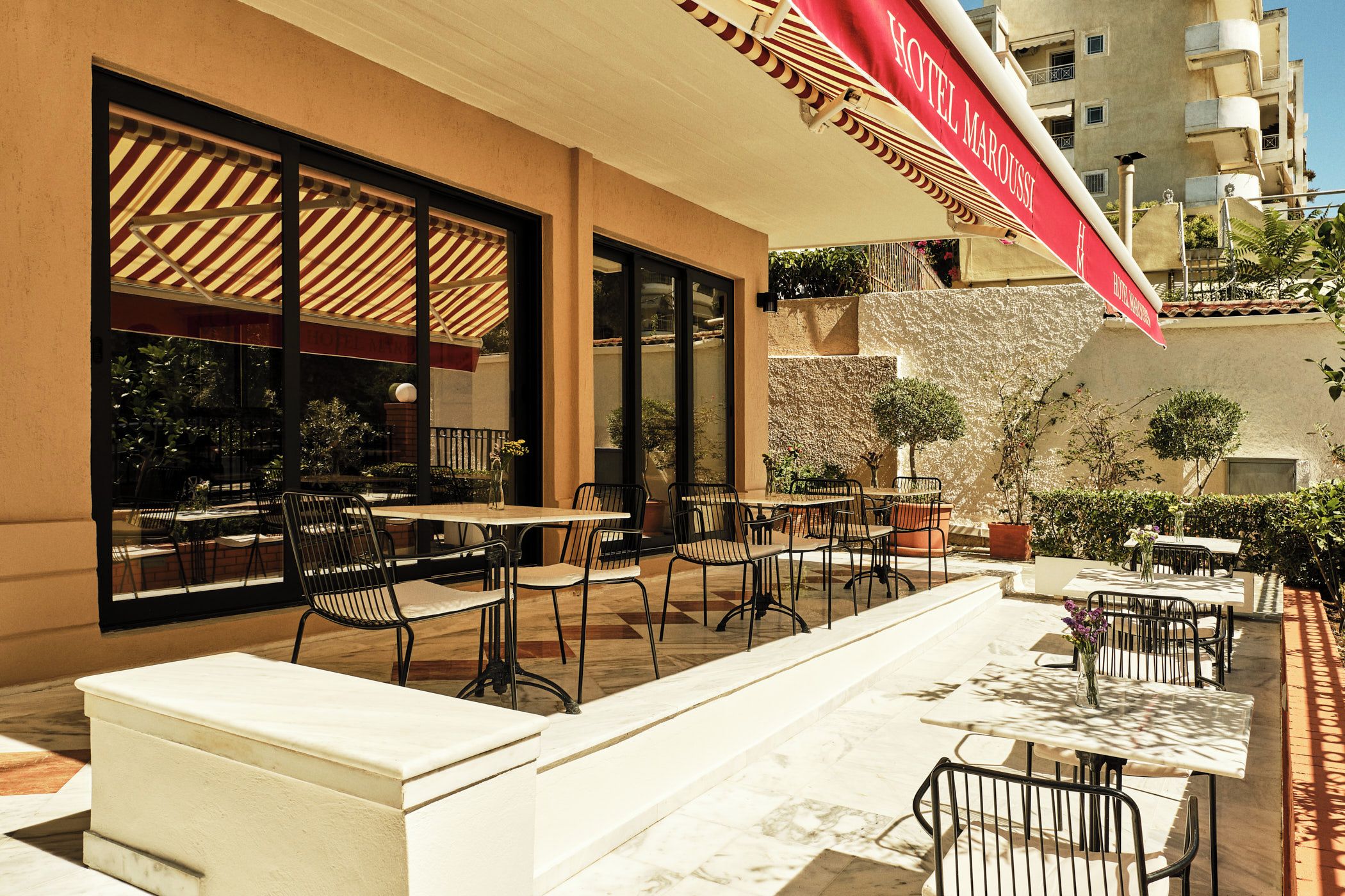 An outdoor seating area with tables and chairs, a red awning with the hotel name, and a stone wall.
