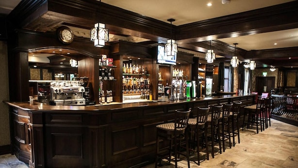 A bar area with a dark wood bar counter, multiple stools, a clock, and a television screen. - Dillons Hotel (Letterkenny)