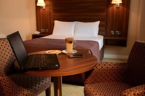 A hotel room with a bed, a bedside table with a lamp, a laptop, a glass of coffee, and patterned armchairs. - Dillons Hotel (Letterkenny)