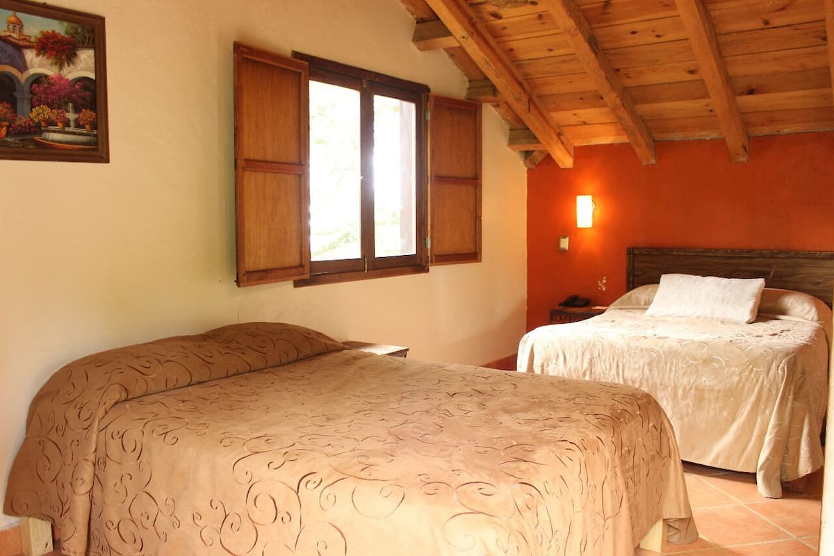 A bedroom with two beds, wooden ceiling beams, a painting on the wall, and a nightstand with a lamp.