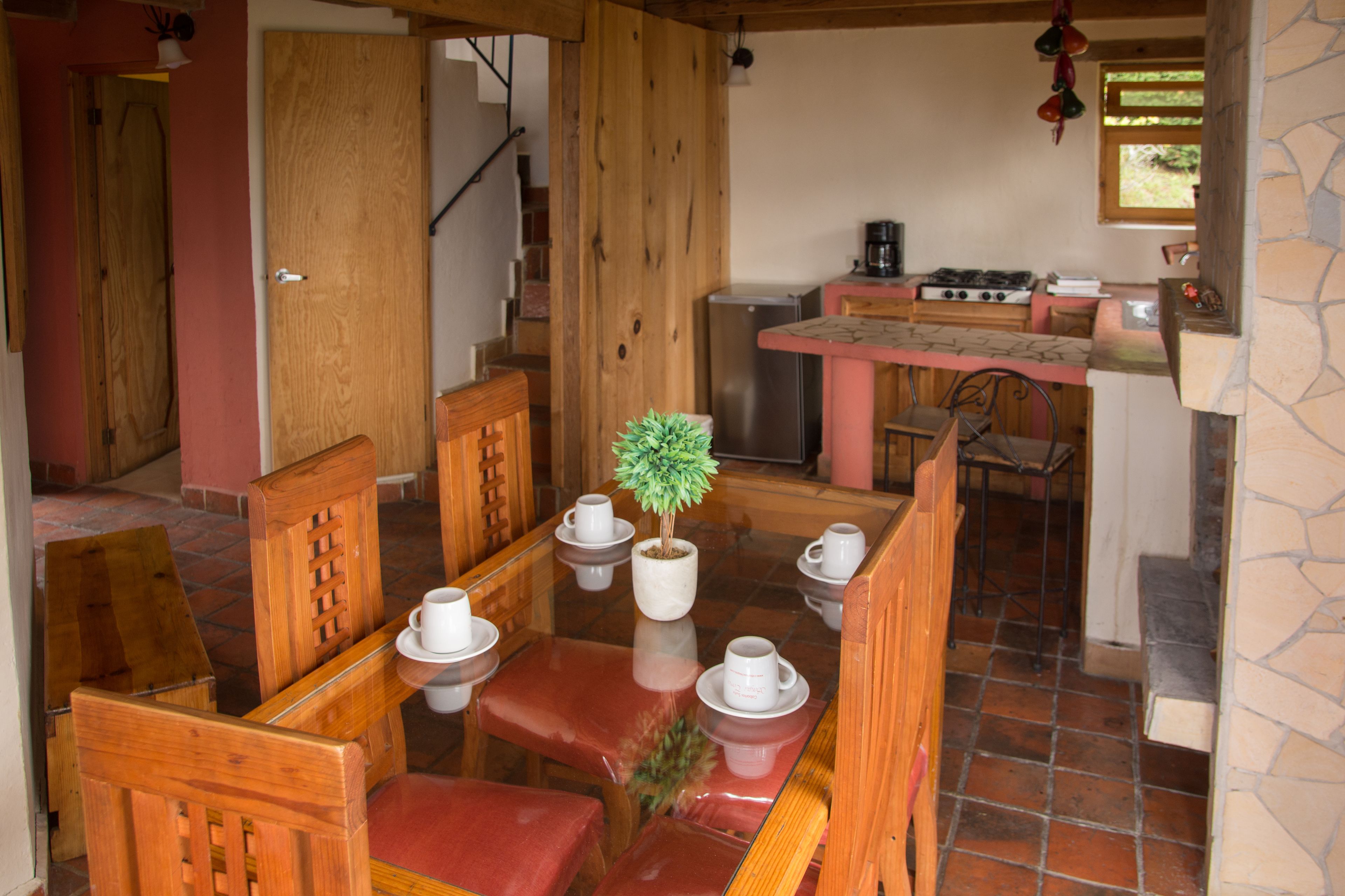 A dining area with wooden furniture, a central table with a potted plant, and a kitchen in the background.