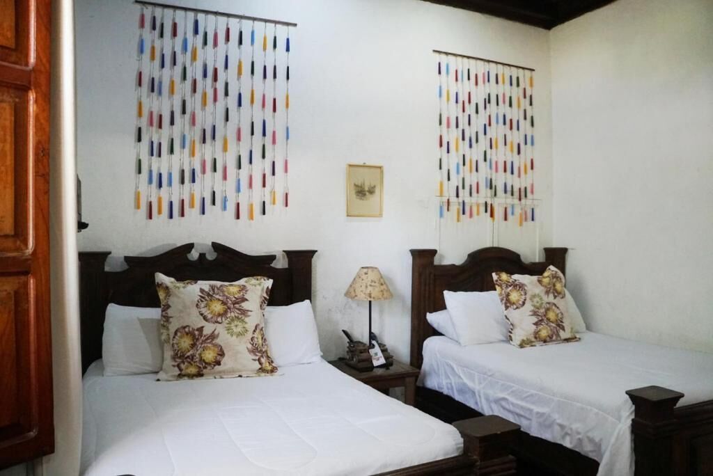 A hotel room with two beds, each with floral-patterned pillows and a wooden headboard. There is a small table with a lamp between the beds, and a framed picture on the wall.