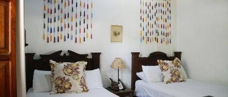 A hotel room with two beds, each with floral-patterned pillows and a wooden headboard. There is a small table with a lamp between the beds, and a framed picture on the wall.