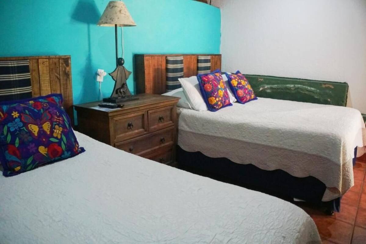 Une chambre avec deux lits, une commode en bois, une lampe et un coussin coloré.