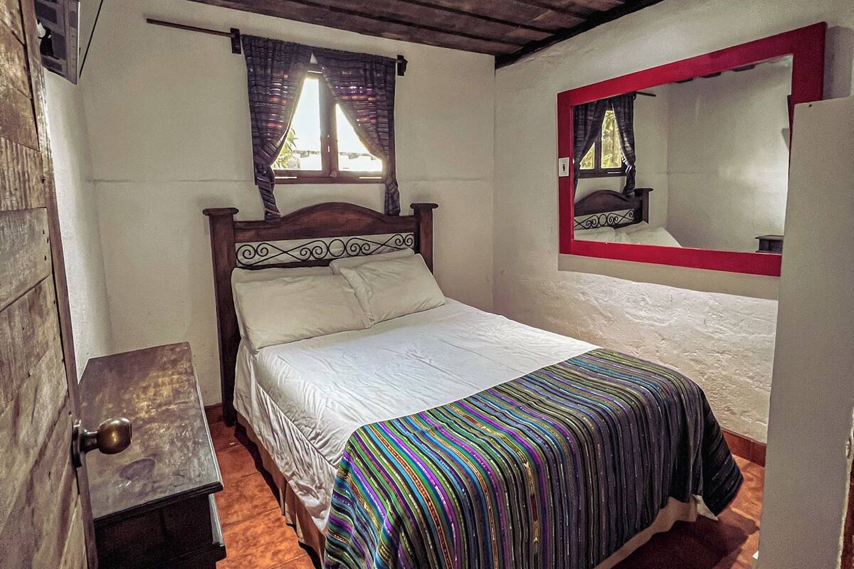 Une chambre avec un plafond en bois, un lit recouvert d’une couverture rayée aux couleurs vives, une petite table de chevet et une fenêtre avec des rideaux.