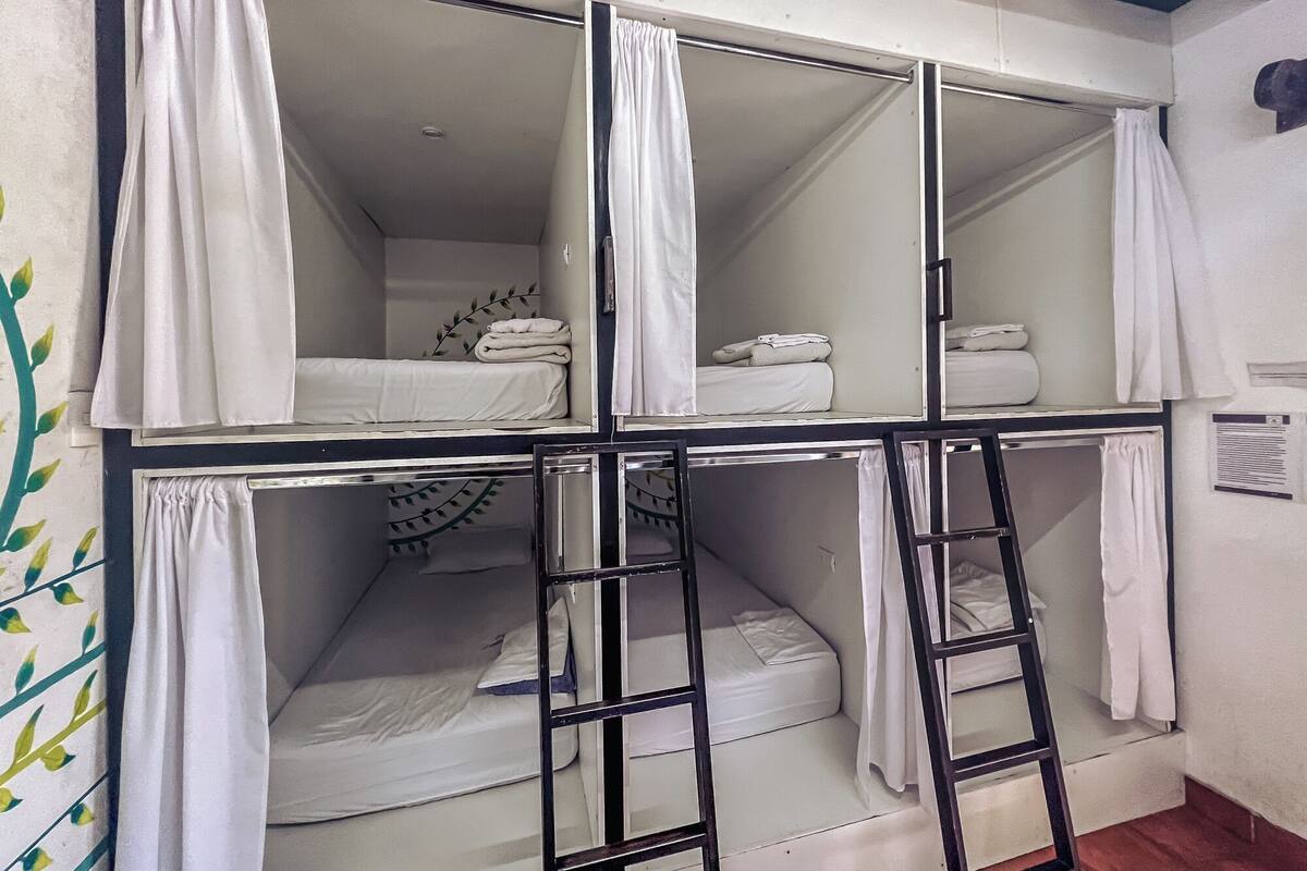 Une chambre avec un lit superposé, du linge de lit et des rideaux blancs, une échelle et un élément décoratif mural.