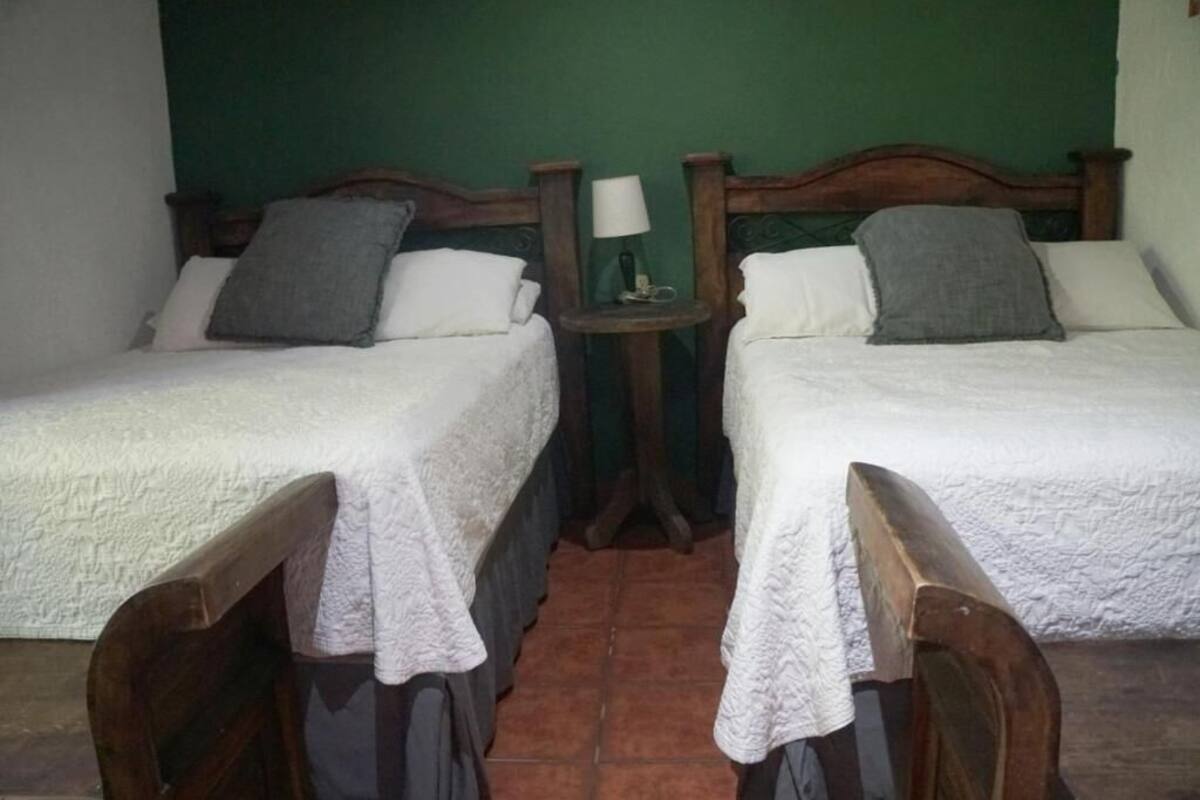 Une chambre avec deux lits en bois, chacun recouvert d’une courtepointe blanche et agrémenté d’un oreiller gris. Une petite table avec une lampe se trouve entre les lits.