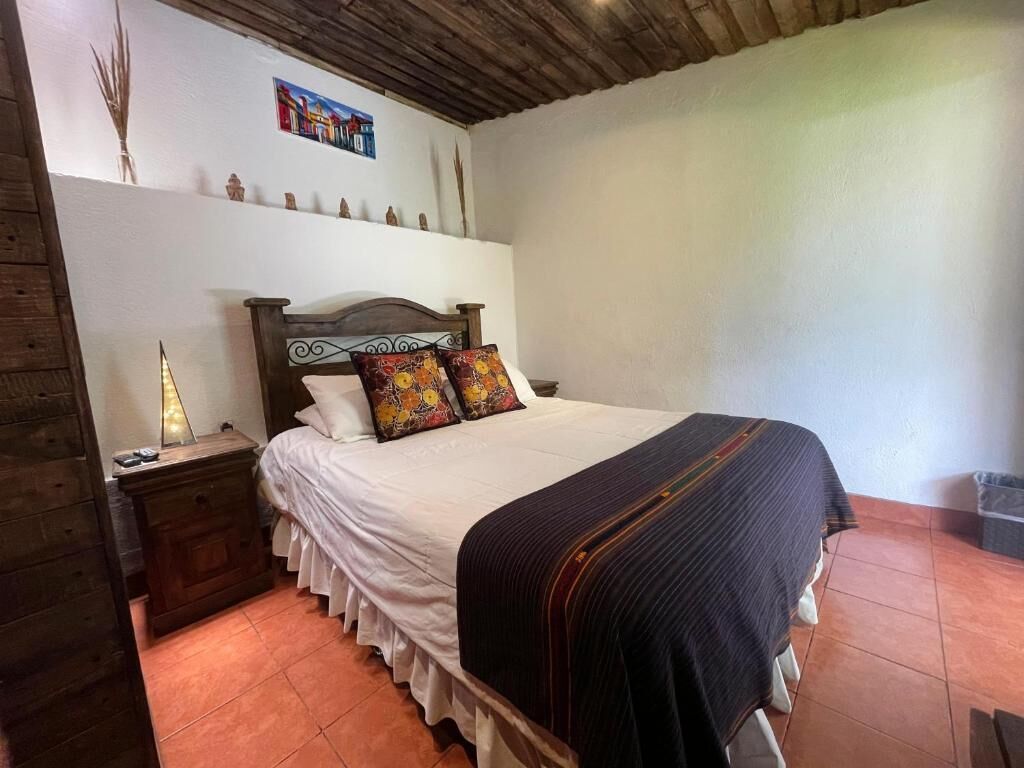 A bedroom with a wooden headboard, a white bedspread, two colorful pillows, a dark blanket, a bedside table with a lamp, and a wall-mounted shelf with decorative items.