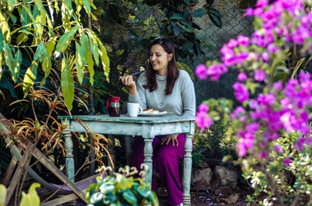 A person sitting at a table with a cup and saucer, surrounded by greenery and flowers.