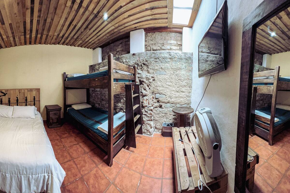 Une chambre de dortoir avec un lit superposé, un lit simple, un mur en pierre et un ventilateur.