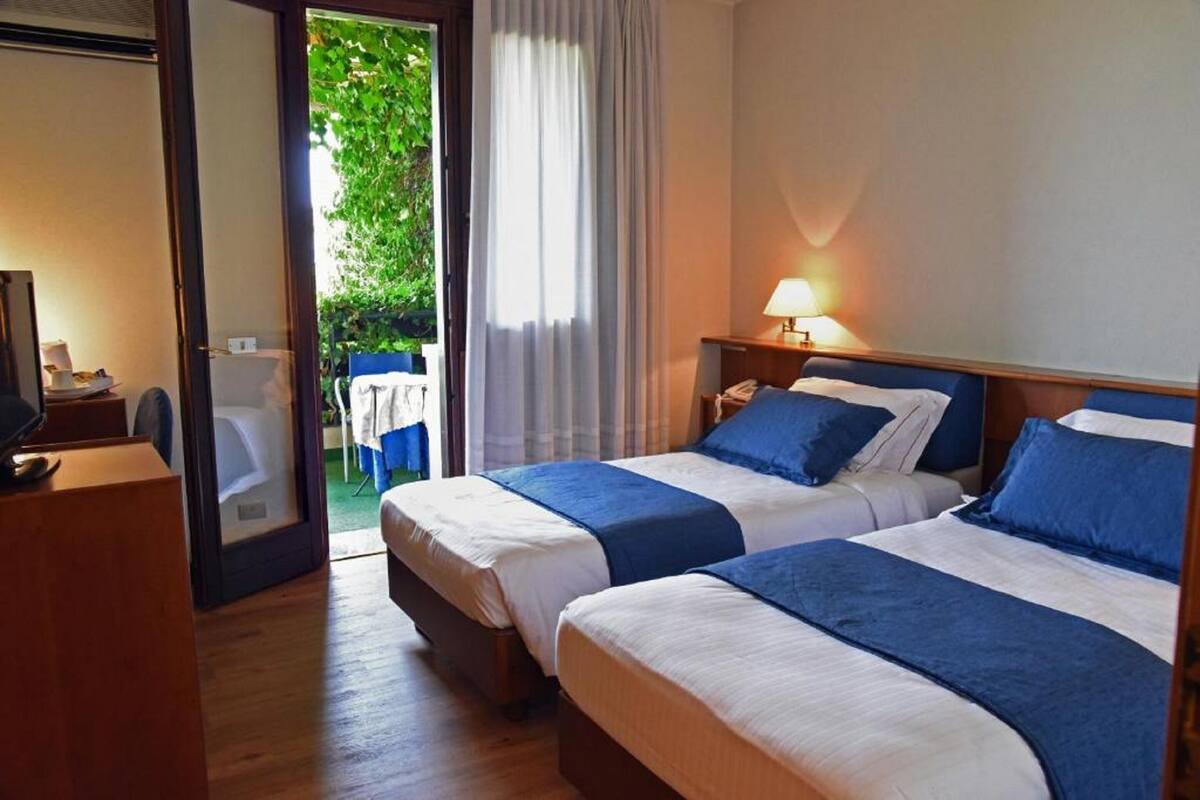 A hotel room with two beds, a desk with a lamp, and a view of greenery through a glass door.