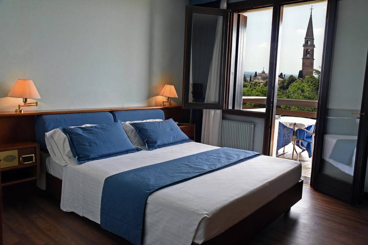 A hotel room with a large bed, two bedside lamps, a view of a church through a glass door, and a balcony with a chair.