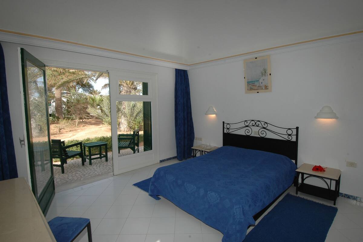 A bedroom with a bed, a desk, and a chair. There is a sliding glass door leading to an outdoor area with a table and chairs.