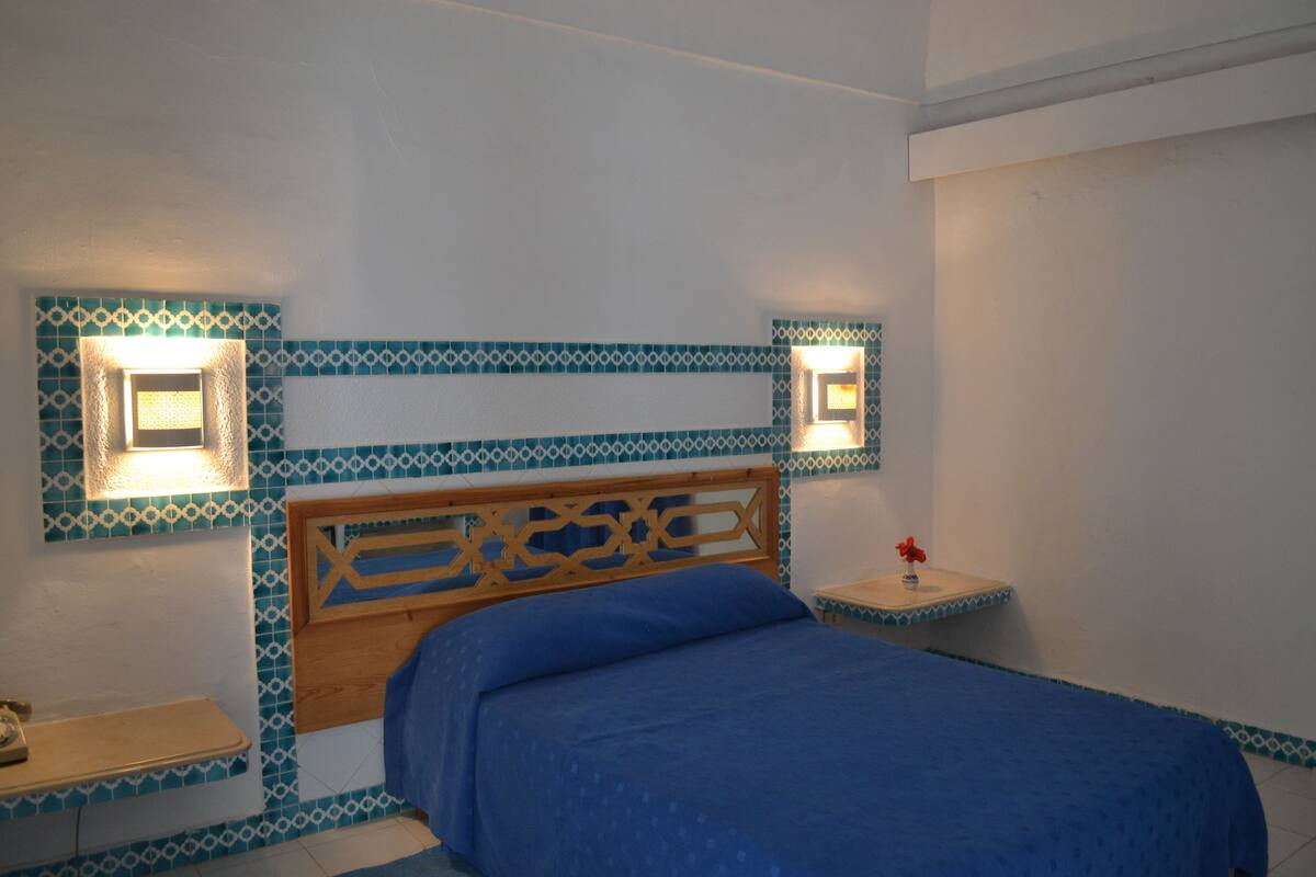 A bedroom with a wooden headboard, a blue bedspread, and patterned wall tiles.