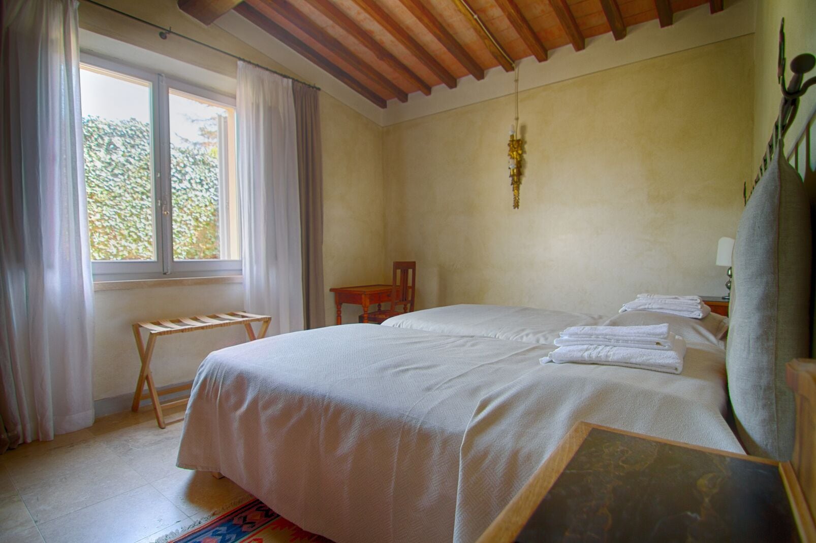 Un dormitorio con una cama grande, una mesita, una silla y un ventanal con cortinas.