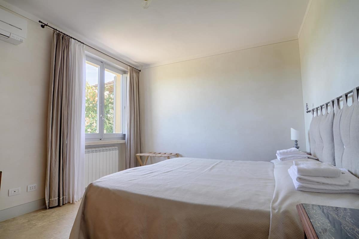 A hotel room with a large bed, a window with curtains, a radiator, and a bedside table with towels.