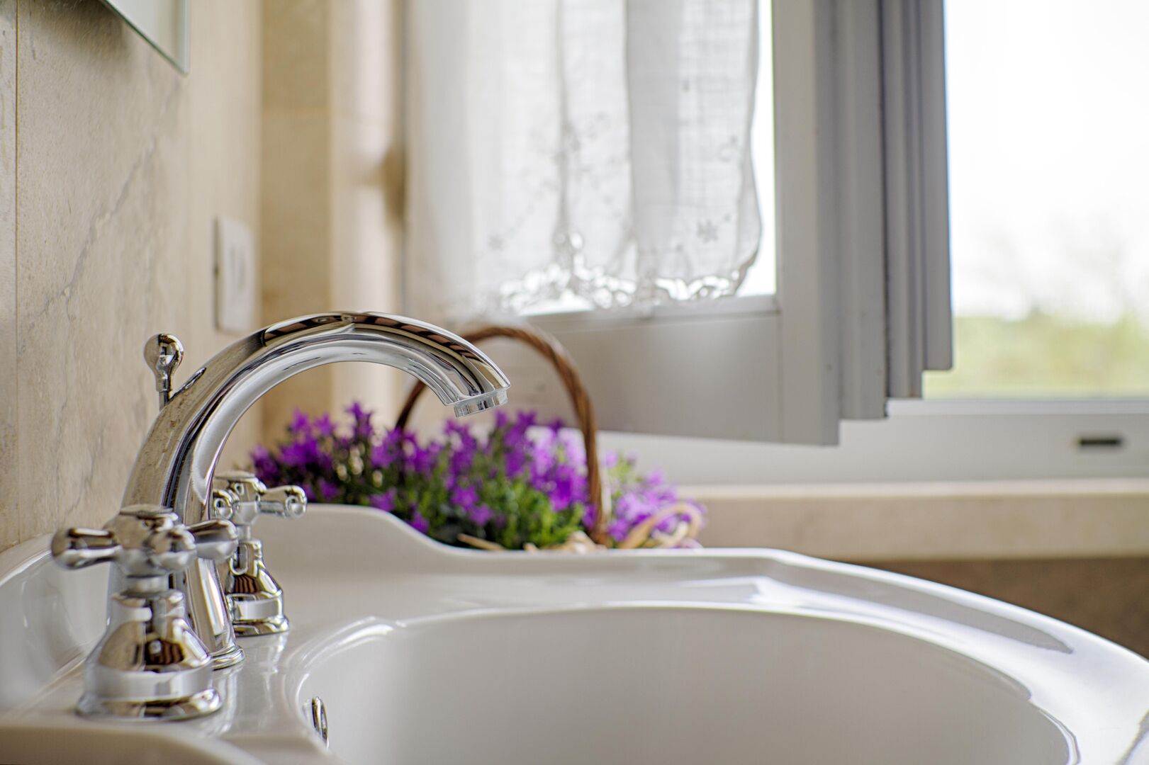 Ein Badezimmer mit weißem Waschbecken, chromfarbenem Wasserhahn und einem Fenster mit Blick auf violette Blumen.