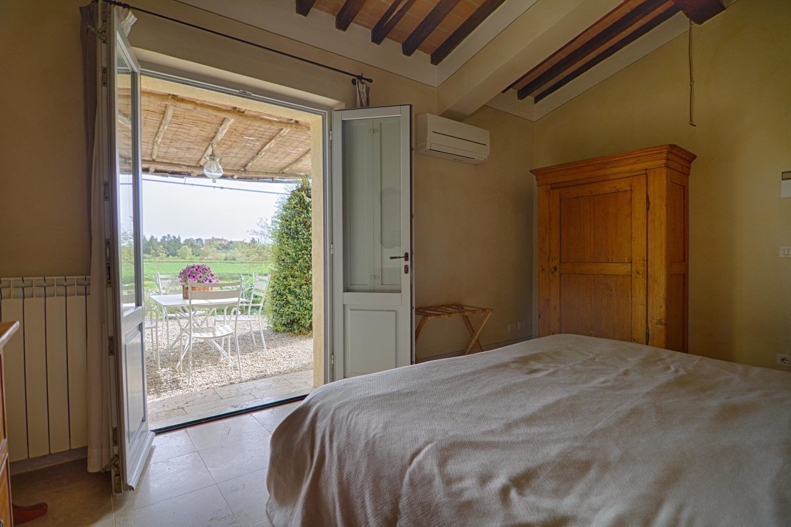 Quarto com uma cama, porta aberta para um pátio com mesa e cadeiras, e um armário de madeira.