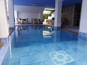 A clear blue swimming pool with tiled flooring, surrounded by wooden chairs and tables under a covered area. - B&B Villa Betta (Bonifati)