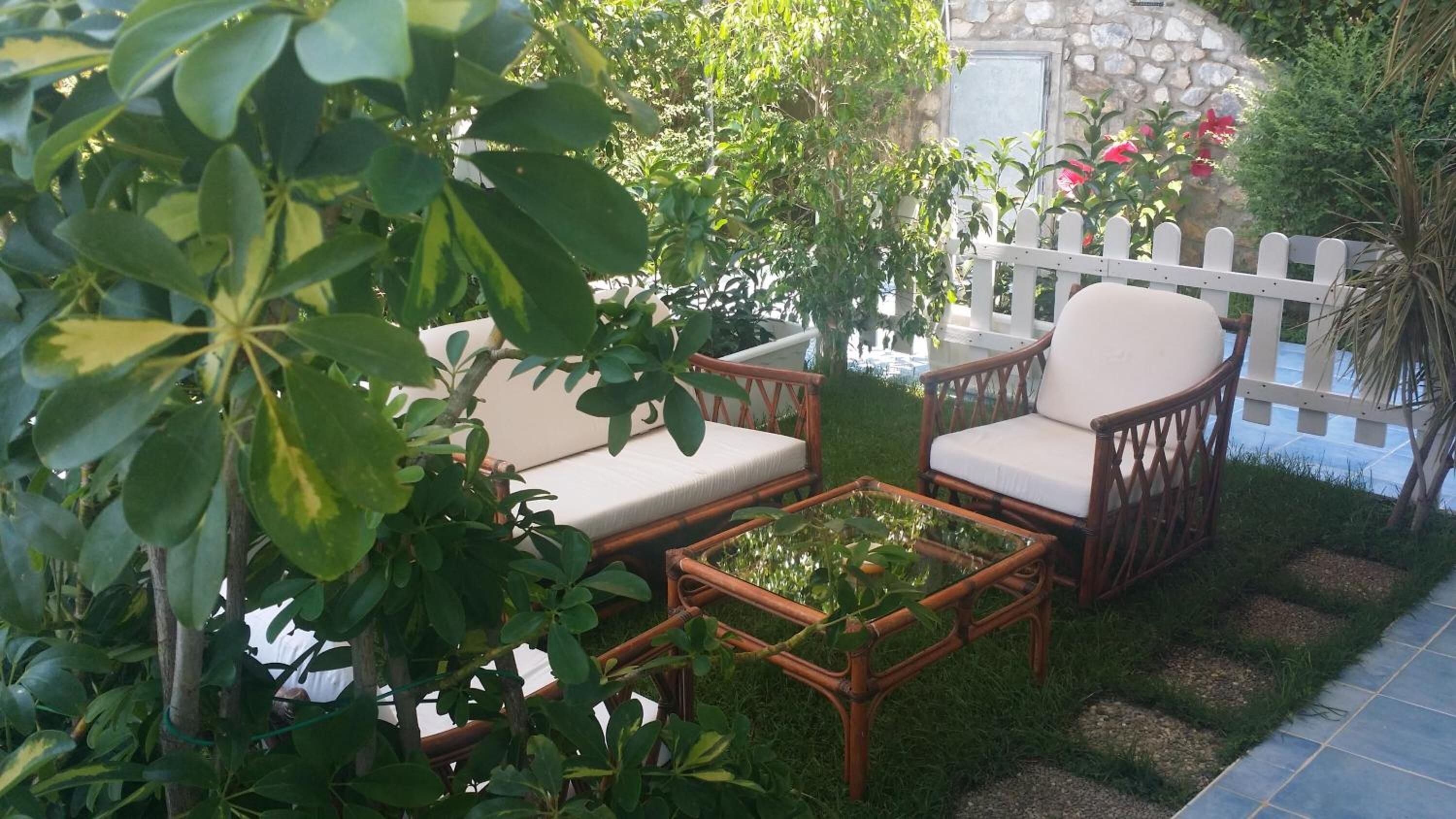 A garden patio with wicker furniture, a white fence, and lush greenery.