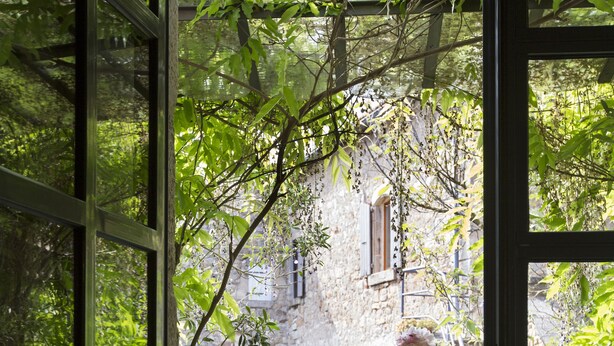 Un balcón con una mesa y una silla, un jarrón de flores y vistas a un edificio de piedra y follaje.