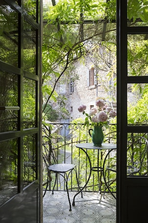 A balcony with a table and chair, a vase of flowers, and a view of a stone building and greenery.