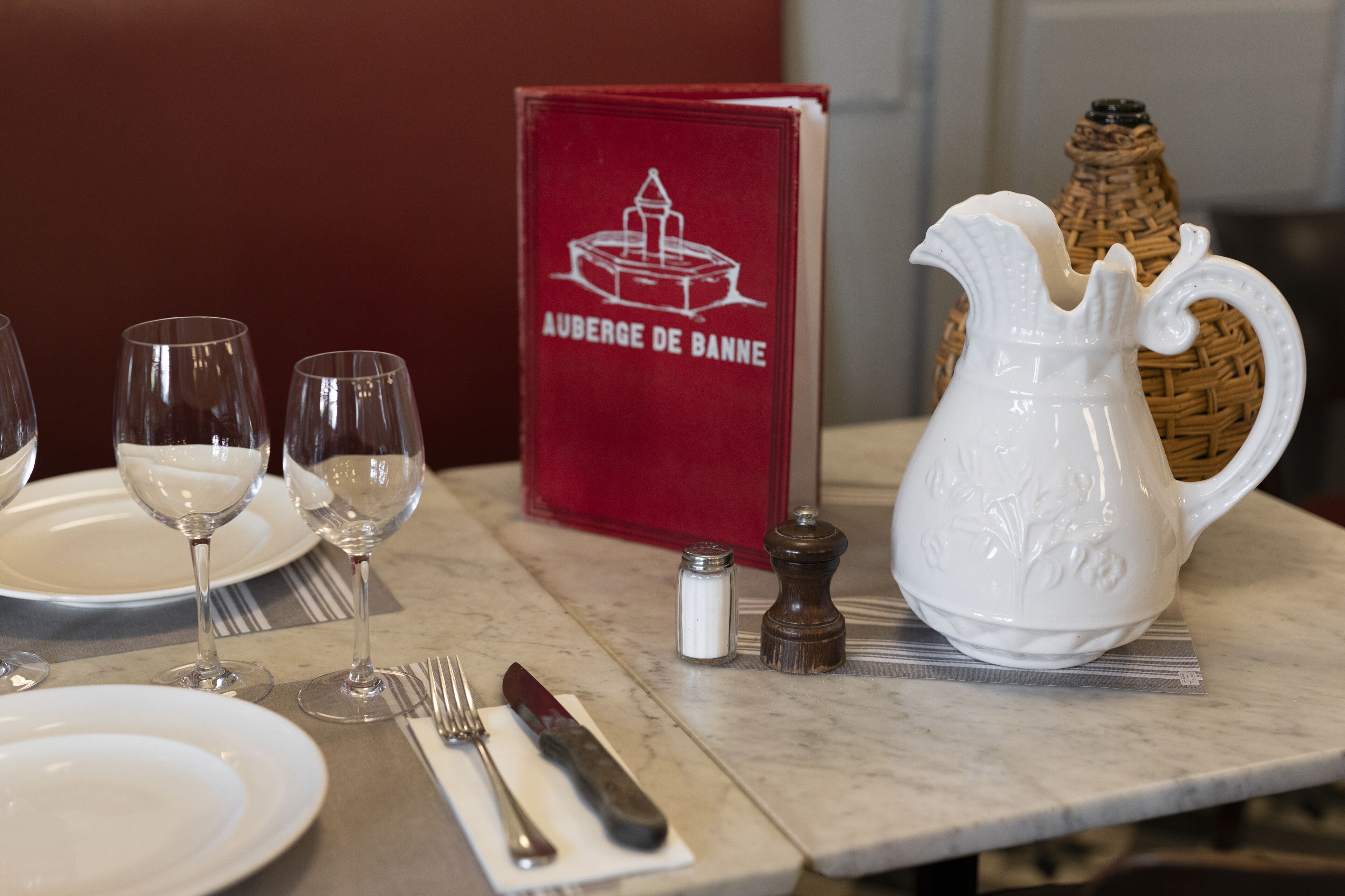 A table set with white plates, wine glasses, a white pitcher, a salt and pepper set, a knife, and a fork on a marble table. A red menu titled "AUBERGE DE BANNÉ" is visible in the background.