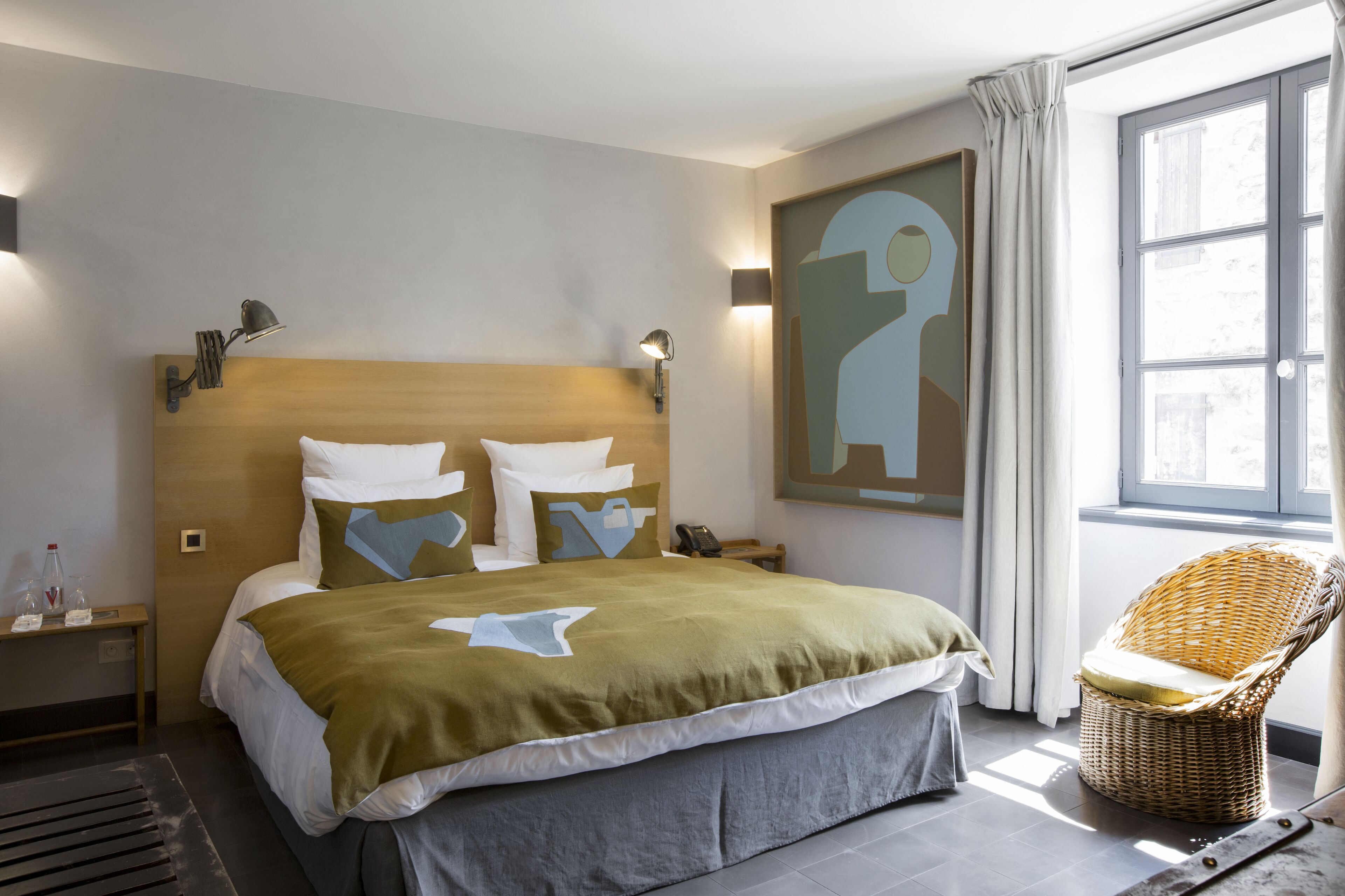 A modern hotel room with a large bed, a wicker chair, and a painting on the wall.