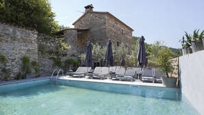 Una piscina cristalina con sillones de descanso y sombrillas, un muro de piedra y un edificio al fondo.