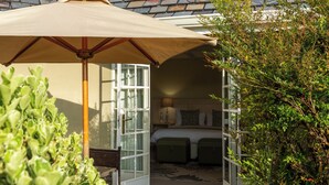 An open doorway to a bedroom with a bed, two nightstands, and a lamp. There is a beige umbrella and a chair outside the doorway. - Moorcroft Manor Boutique Country Hotel (Himeville)