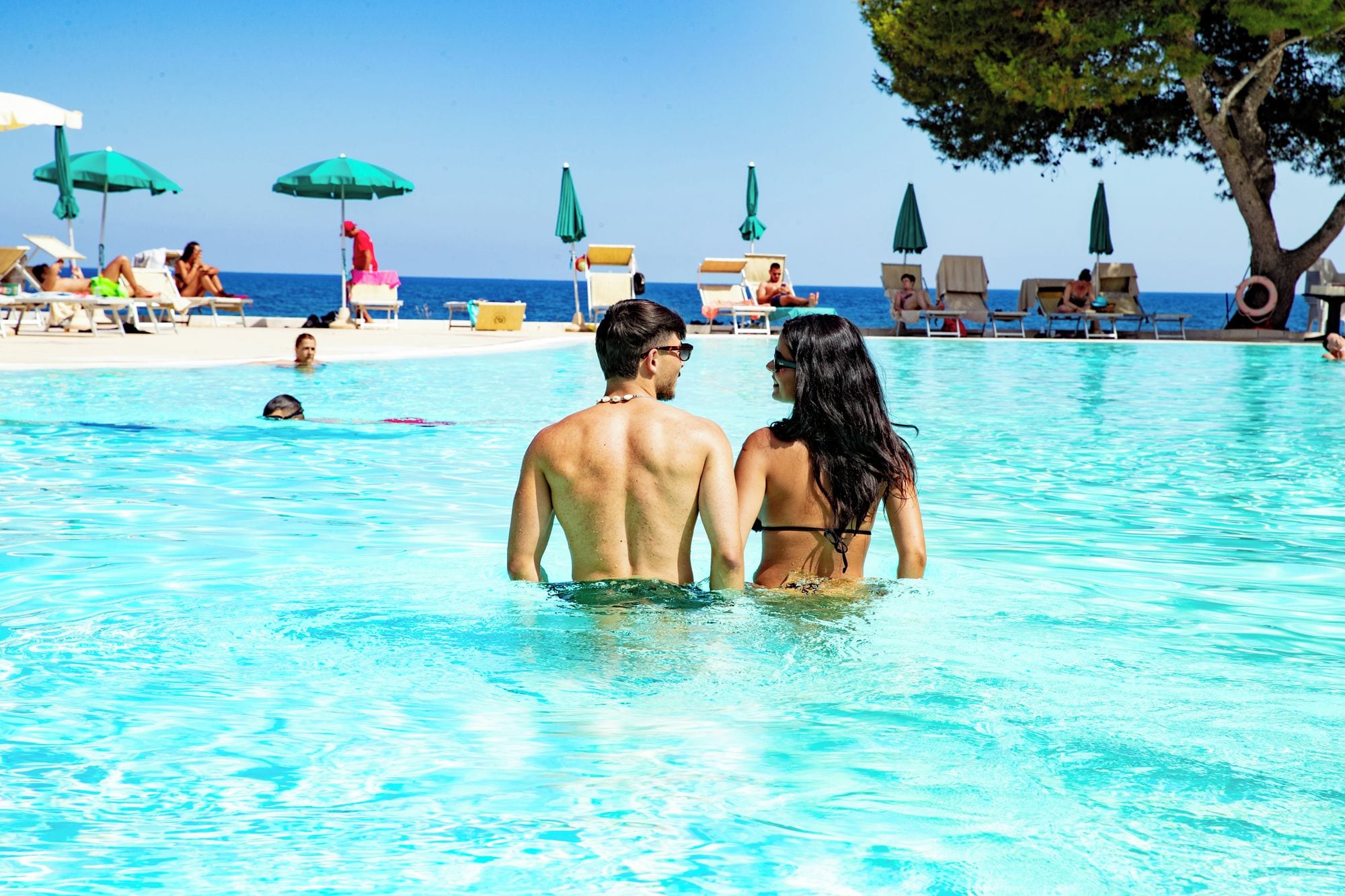 A couple standing in a pool with lounge chairs and umbrellas in the background.