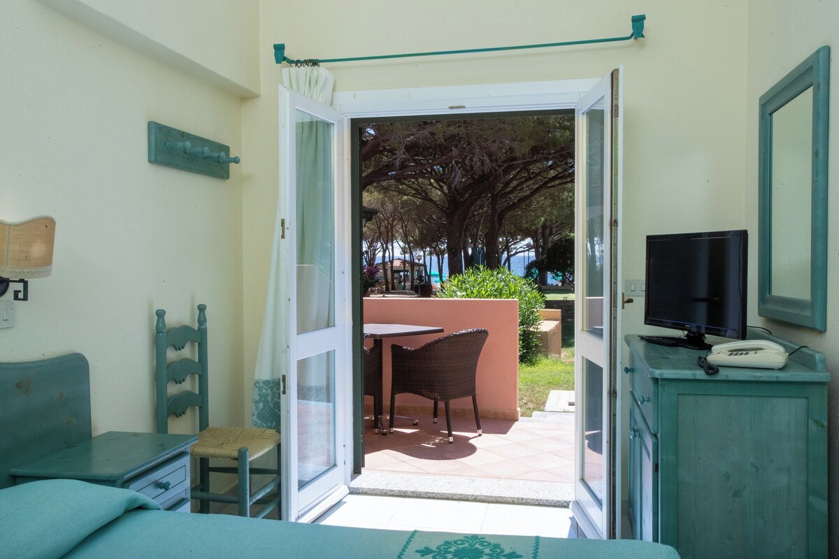 A room with a bed, a desk, a chair, a TV, and a phone. There is a door leading to an outdoor area with a table and chairs.