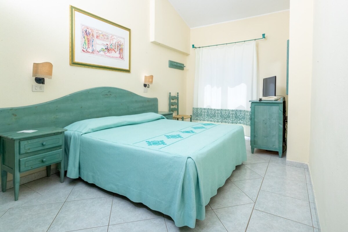 A bedroom with a bed, a nightstand, a dresser, a television, and a wall-mounted light fixture.