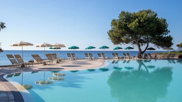 Een zwembadterras met ligbedden en parasols, met vrij uitzicht op zee, en een boom die schaduw biedt.