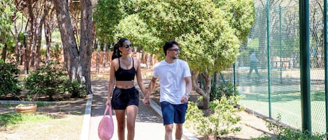 Two people walking on a paved path in a park with trees and a fence in the background.