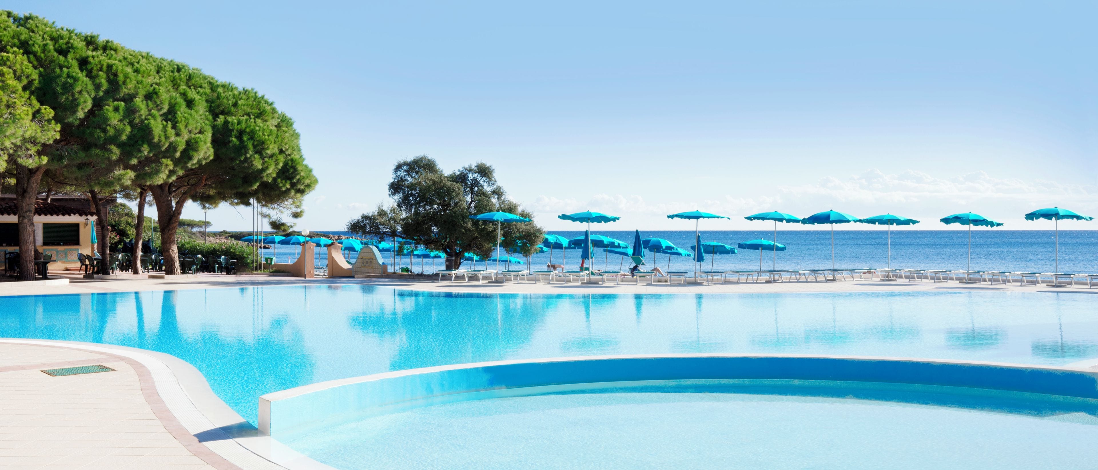 A large swimming pool with blue umbrellas and lounge chairs, surrounded by trees and a view of the sea.