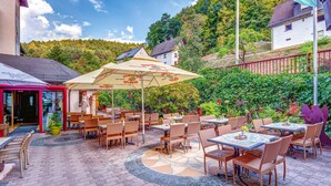 An outdoor cafe with wicker furniture, a large umbrella, and a view of a hillside. - Hotel Christel (Heimbuchenthal)