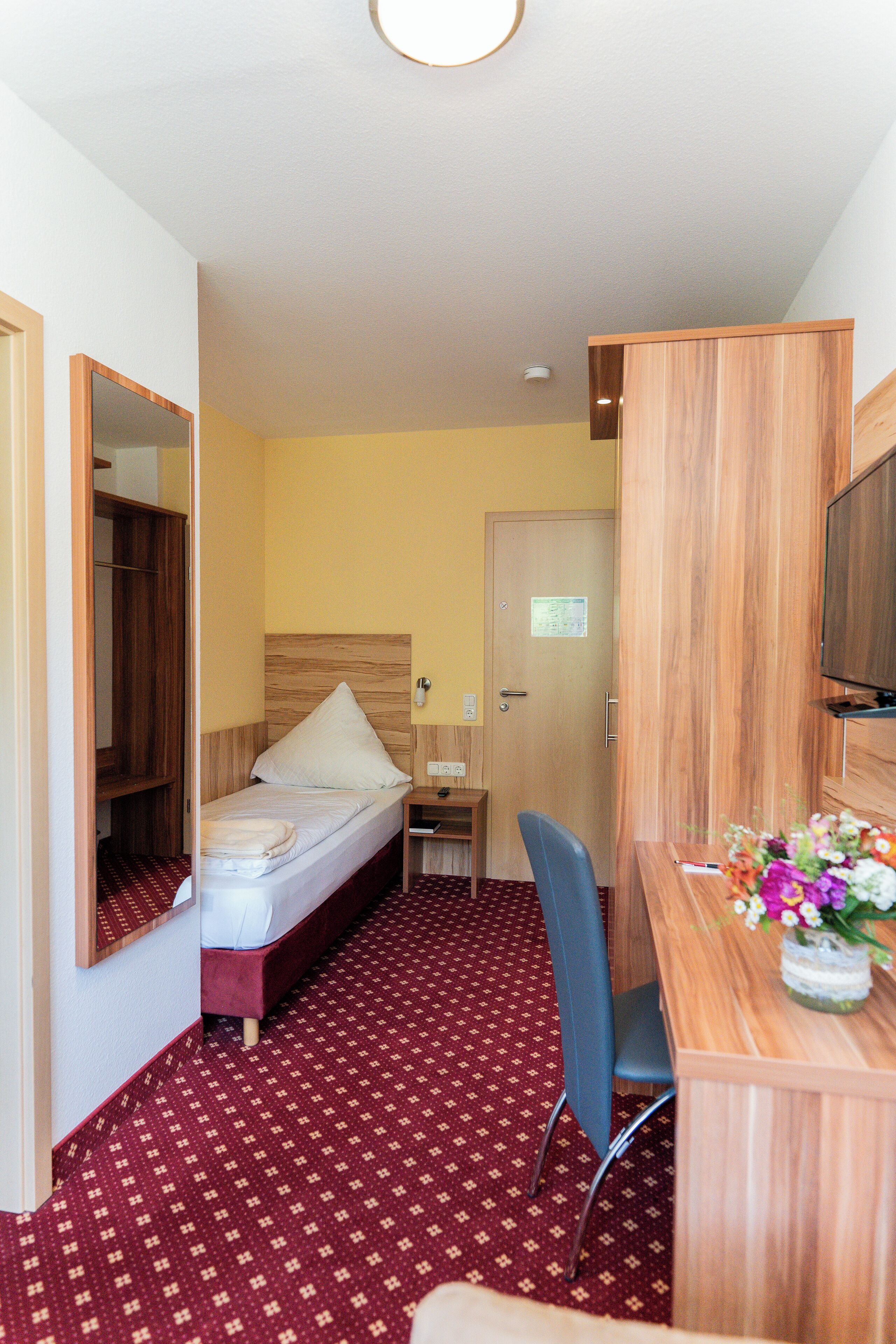 A hotel room with a single bed, a desk with a chair, a mirror, and a TV.