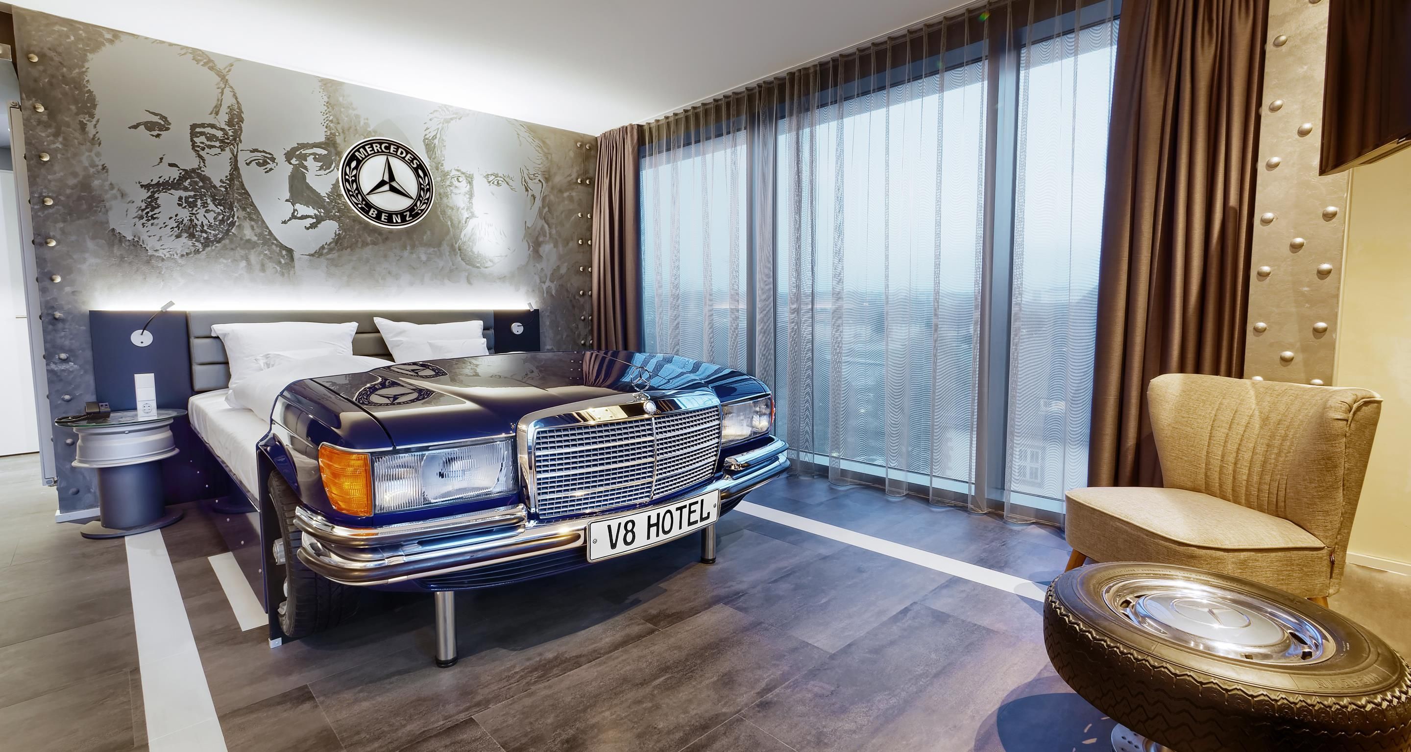 A hotel room with a bed, a large window, a wall with a Mercedes-Benz logo, and a car-shaped headboard.