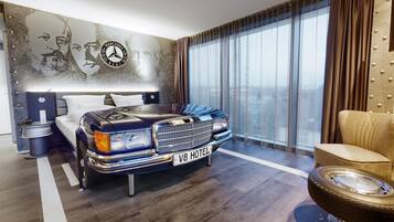 A hotel room with a bed, a large window, a wall with a Mercedes-Benz logo, and a car-shaped headboard.