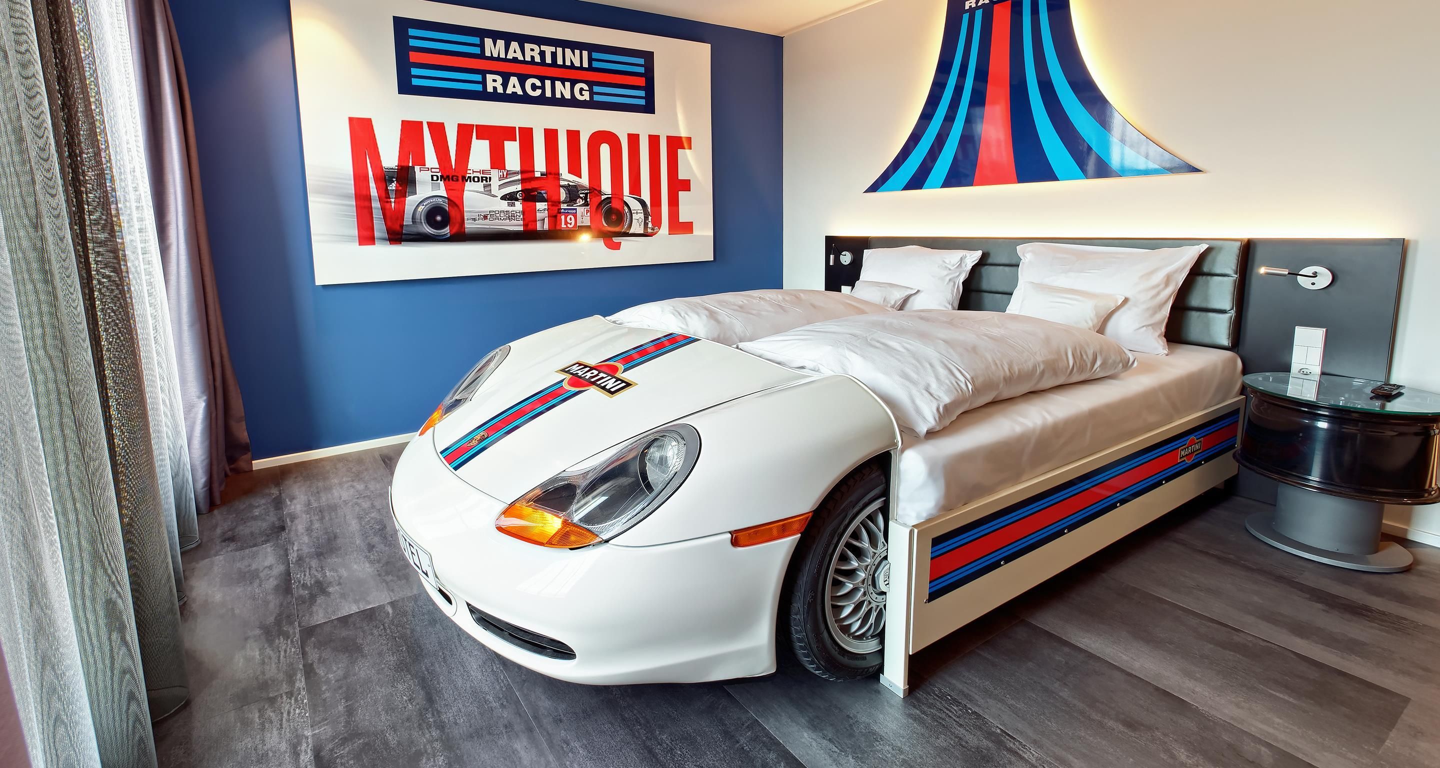 A bedroom with a bed featuring a racing car design, a wall with a racing theme, and a modern lamp.
